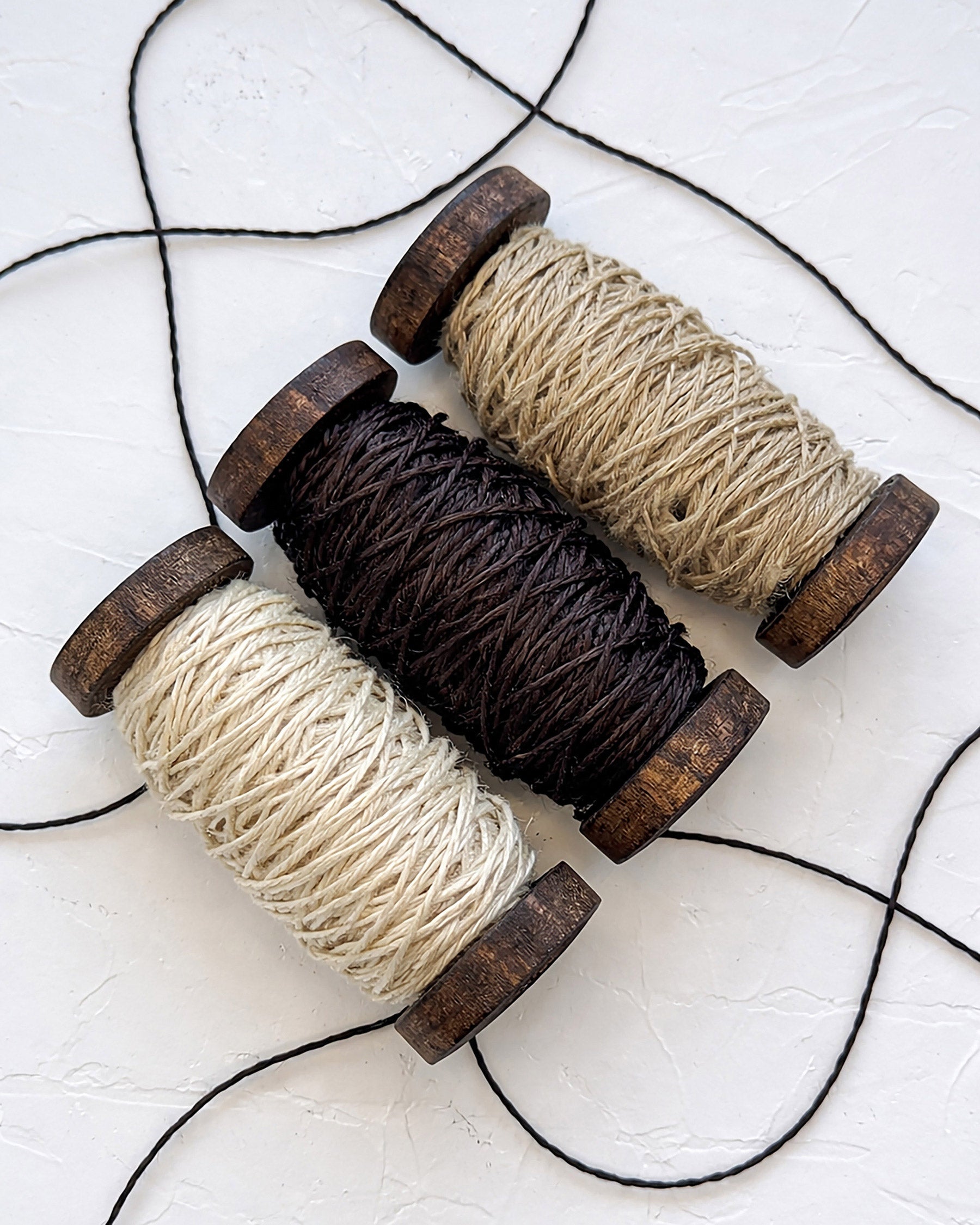 Three spools of linen twine thread in ecru, dark chocolate, and natural flax color on a white surface